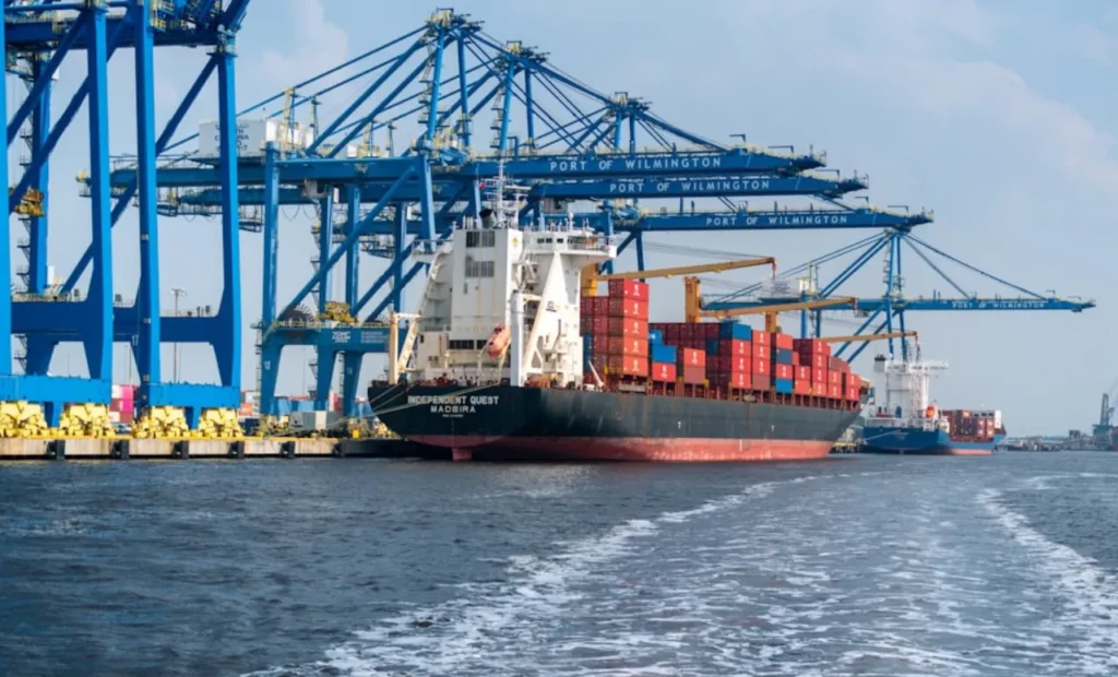 A large cargo ship docked at a port with blue cranes and containers.